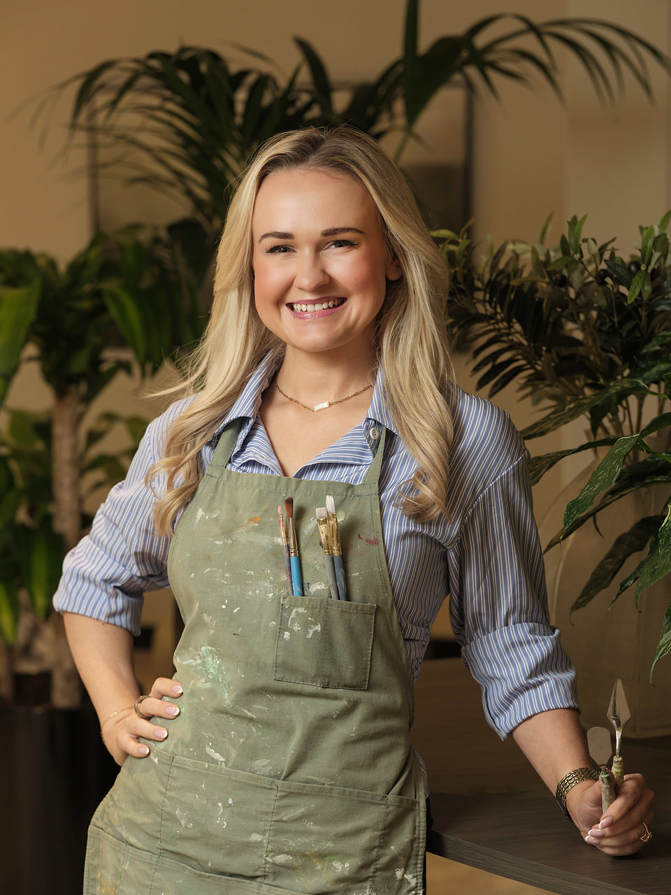 Madie Brunt smiling in a paint-splattered green apron with brushes in the pocket, holding palette knives in front of leafy plants