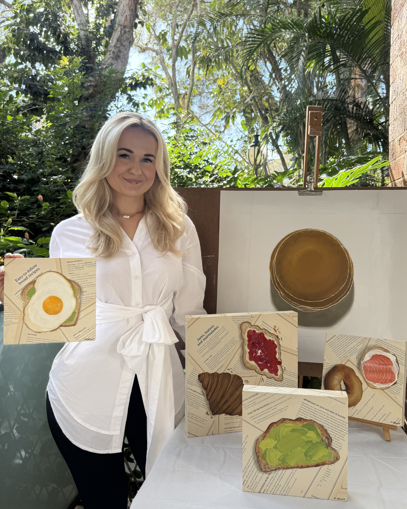 Madie Brunt holding one of her food art paintings, surrounded by her artwork in a garden setting